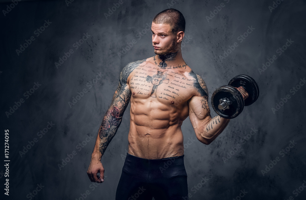 Shirtless, tattooed male holds dumbbell over grey artistic background.