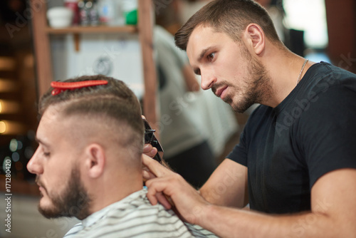 Handsome young male barber giving his client a haircut using a clipper working at his barbershop.