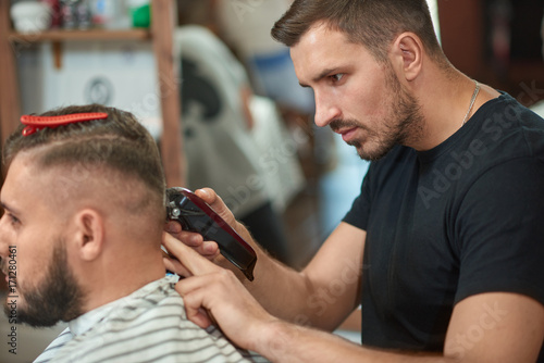 Bearded young male barber cutting hair of his male client with a clipper while working at his barbershop.