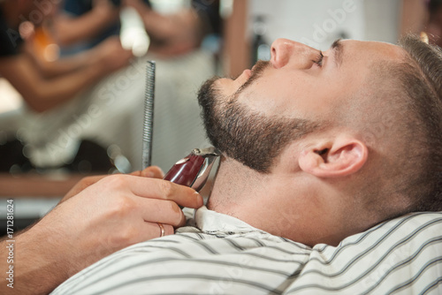Close up portrait of a handsome young bearded man getting his beard trimmed by a professional barber. 