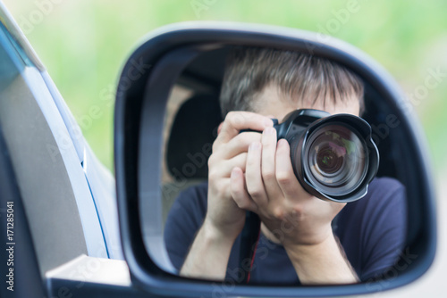 A man is taking photo someone or something from an open car window. Reflection in the side mirror of the car