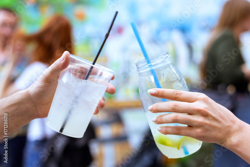 Drinking lemonade cocktail glasses with straw and ice shaking, man and woman friend hands cheers at an event with blurred red hair people and colored wall background