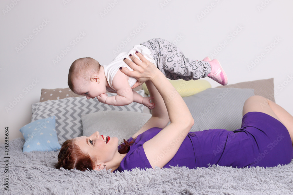 Young mother raises her little baby on bed with pillows in bedroom Young mother raises her little baby on bed with pillows in bedroom