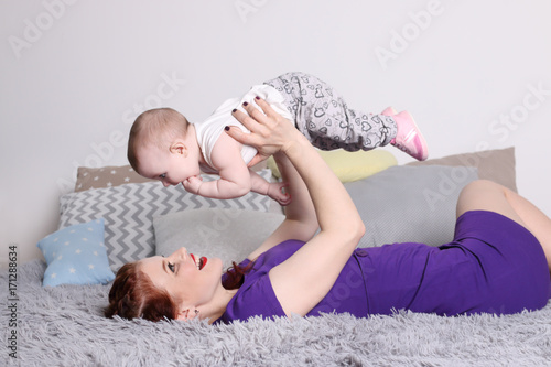 Young mother raises her little baby on bed with pillows in bedroom