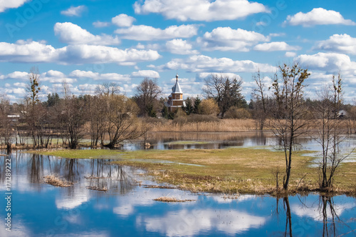 Spring high water flooded agricultural fields and trees due to flood of river against background of the village and of Orthodox Church with reflection clouds and blue sky in water in temperate zone