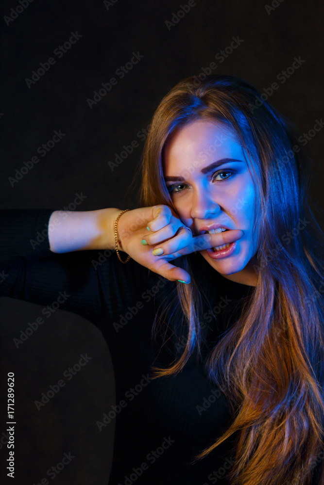 Blonde in black bites her finger in black studio with blue light Blonde in black bites her finger in black studio with blue light