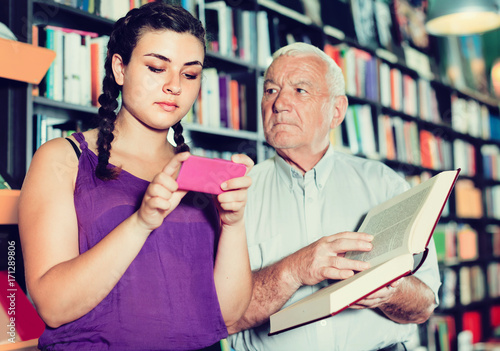 Old man is choosing book while girl chatting by phone in bookstore.