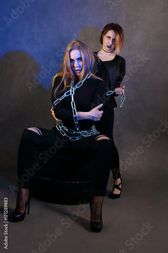 Two girl in black poses on tires with chain in studio with blue light, focus on blonde