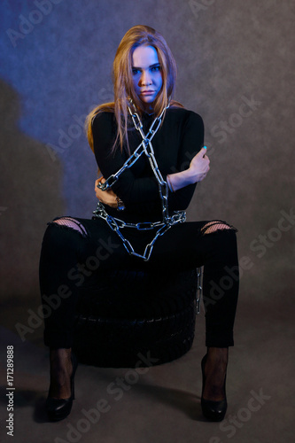 Girl in black clothes in chain poses on tires in studio with blue light