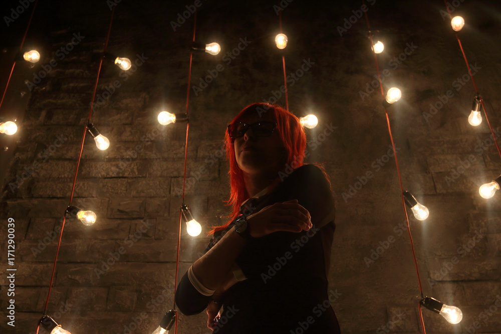Fototapeta premium Girl in glasses poses near wall in black studio with garlands of lamps, under view