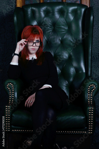 Woman in glasses and black dress sits in leather armchair in black studio