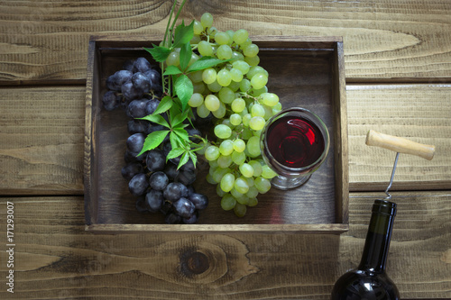 Open bottle of wine with a glass, corkscrew and ripe grape in tray on a wooden background. Copy space and top view.