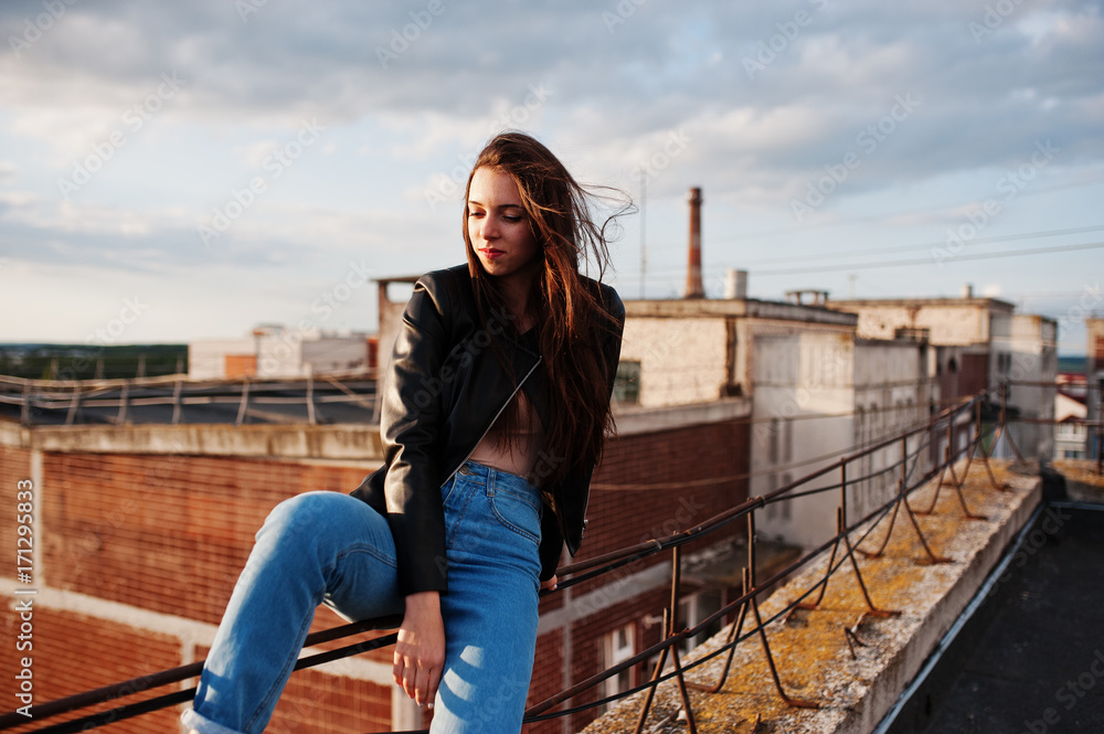 Portrait of a gorgeous young woman in black leather jacket, jeans and sneakers sitting on handrails on the roof with picturesque view of a park.