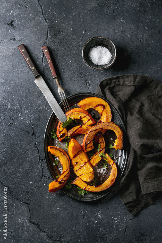 Roasted sliced pumpkin with balsamic sauce, greens and sea salt. Served on vintage metal tray with textile napkin and cutlery over black texture background. Top view with space