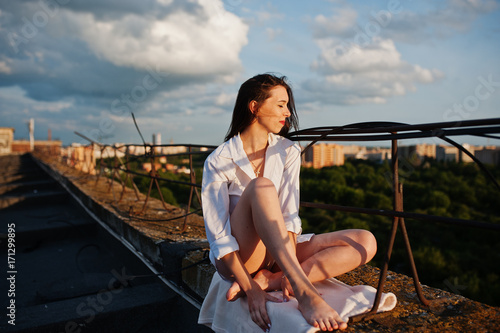 Portrait of a fabulous young woman in white male shirt sitting on the edge of the building roof with a fantastic view of a city in a background.