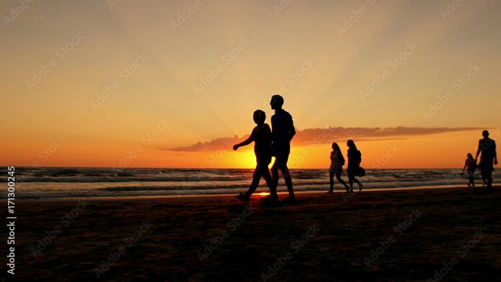 Silhouettes of people walking along the sea beach against the background of sea waves and red sunset. Silhouettes of people walking along the sea beach against the background of sea waves and red sunset.