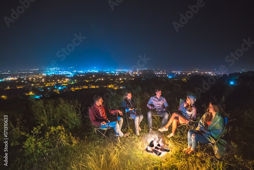 The five people sit near the bonfire on the background of the city. night time
