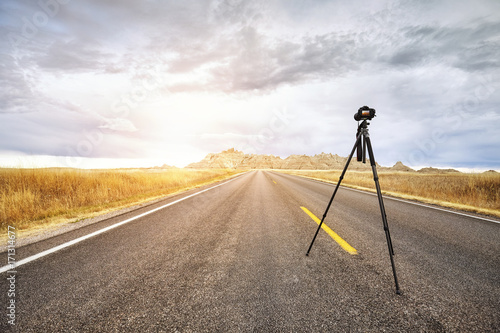 Professional photo camera on tripod on an empty road at sunset, focus on the camera, travel or work concept, Badlands National Park, South Dakota, USA.