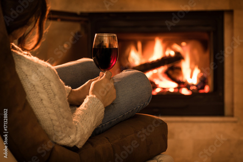 Young woman sitting at home by the fireplace and drinking a red wine.