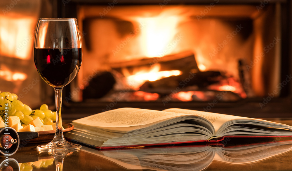 Naklejka premium Red wine glass with an open book on table in front of burning fireplace.