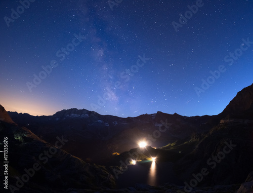 The colorful glowing Milky Way arch and the starry sky from high up on the Alps. Lights from hydroelectric lake dam.