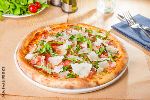 Pizza with prosciutto, parma ham, arugula and parmesan on served wooden restaurant table. Italian cuisine. Selective focus