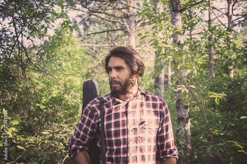 handsome male with beard and long hair walking in the summer forest with guitar case   © serejkakovalev
