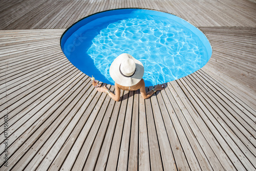 Woman in big sunhat with cocktail drink relaxing at the round swimming pool with blue water outdoors. Top view