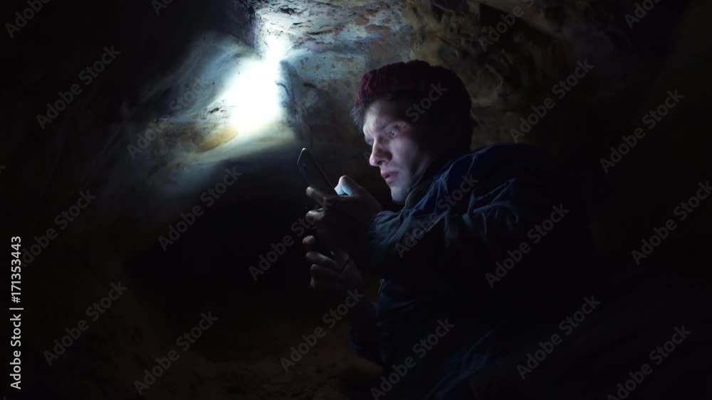 Man using smartphone laying on ground into very narrow cave with hand flashlight Man using smartphone laying on ground into very narrow cave with hand flashlight