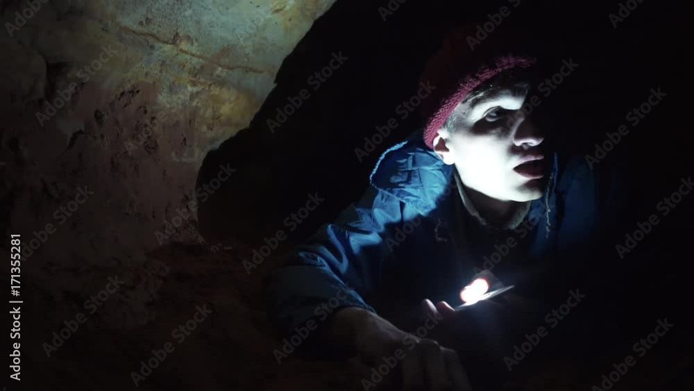 Young explorer scrambles by the ground inside narrow cave illuminates the way with flashlight in his smartphone. Close up shot Young explorer scrambles by the ground inside narrow cave illuminates the way with flashlight in his smartphone. Close up shot