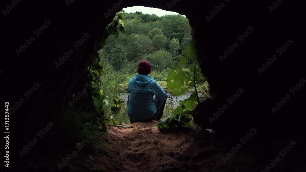 Traveler using smartphone sitting on the ground near the exit from cave. A look from the cave. Deep forest at background. Traveler using smartphone sitting on the ground near the exit from cave. A look from the cave. Deep forest at background.