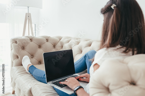 Beautiful young woman working from home on a laptop lying on the sofa in the room, back view