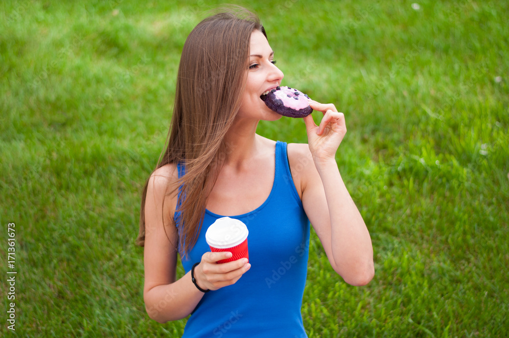 Cheerful young woman taking a bite of the donut and holds a coffee Cup outdoors
