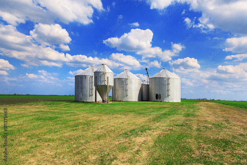 Metal grain bins on farmland in a rural background. Blue and clouds in the background.