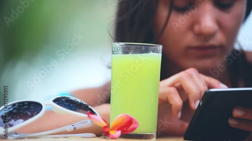 Young attractive woman using mobile phone drinking healthy juice on the beach next to sunglasses and frangipani flower on blurred bokeh background. 1920x1080 photo