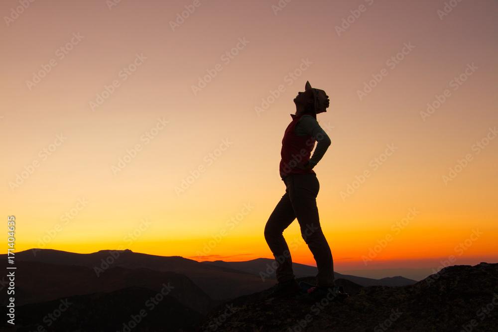 Sunset Silhouette of attractive woman staying and looking happy on top of the mountain. Freedom, adventure and leisure vacation concept.