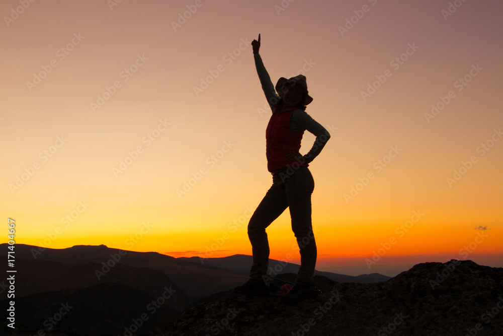 Sunset Silhouette of attractive woman staying and looking happy on top of the mountain. Freedom, adventure and leisure vacation concept.