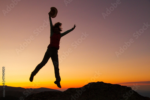 Silhouette of happy joyful young attractive woman jumping and having fun at the mountain against the sunset. Freedom, adventure and leisure vacation concept.