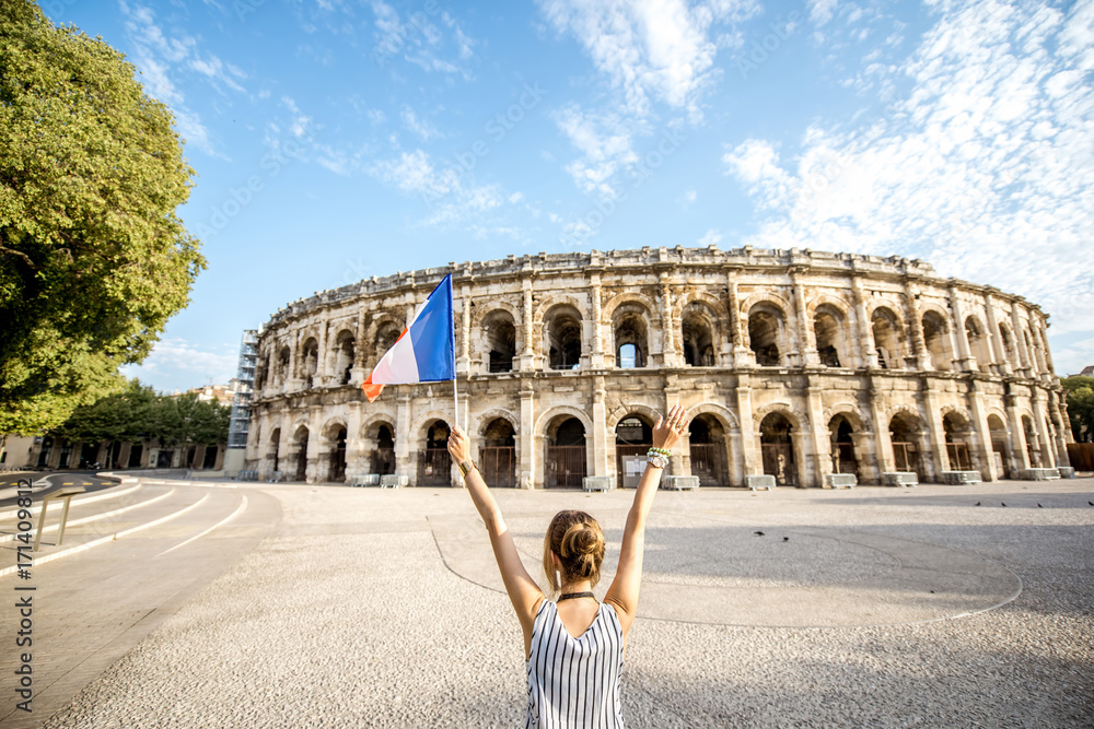 Young woman tourist standing back with french flag in front of the ancient amphitheatre in the old town of Nimes during the sunny morning in France.