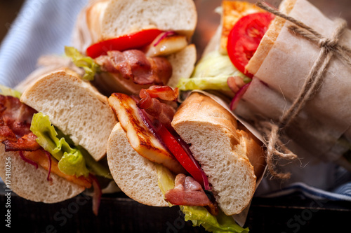 Fresh baguette sandwiches bahn-mi styled with bacon, roasted cheese, tomatoes and lettuce on wooden background. Close up view, horizontal composition.