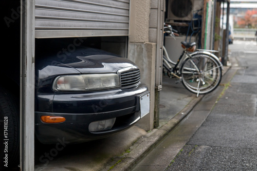 Fototapeta Naklejka Na Ścianę i Meble -  The parked car exceeds the size of a garage in the house, Kyoto, Japan. The car is parked in a small garage in the city center. Front of the car sticking under the door of the garage.