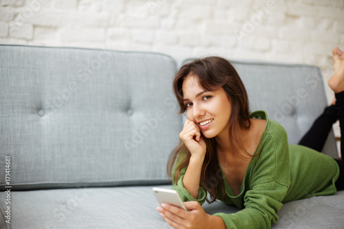 Picture of beautiful young mixed race female with playful charming smile relaxing on comfortable couch at home, texting boyfriend via online messenger using electronic device. People and technology