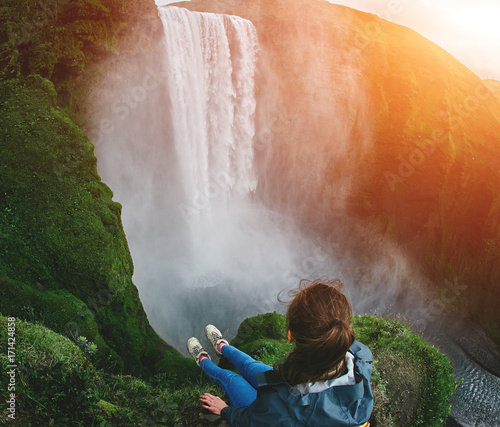 Girl in waterproof clothing sits on the cliff on background of Skogafoss waterfall in Iceland. View from above photo