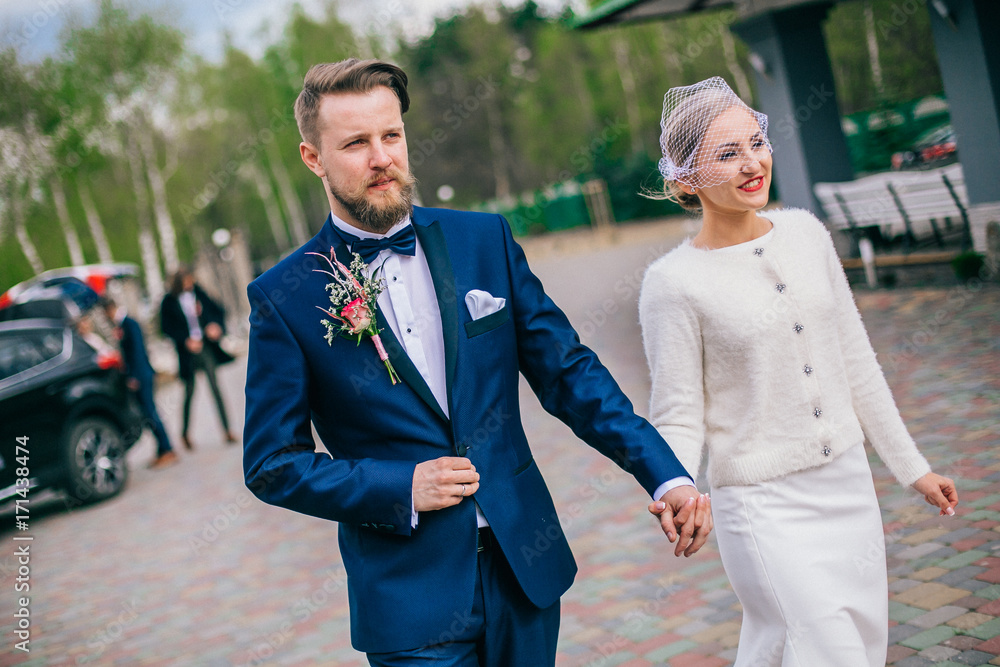 Happy newlywed couple on a walk in old European town street, gorgeous bride in white wedding dress together with handsome groom in stylish suit and beard.