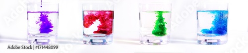 On a white background four glass transparent glasses with water, in each drip multicolored paints. Ink blurs in the water, form beautiful patterns.