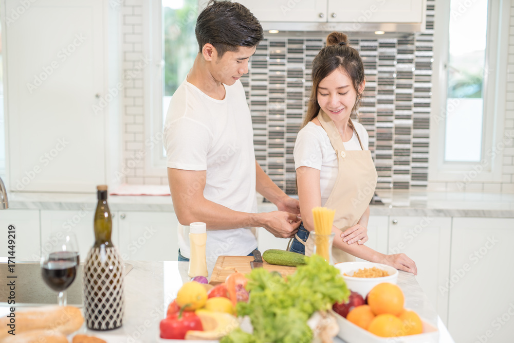 Happy young asian couple, husband help his wife to put on apron before cooking in the kitchen at home.