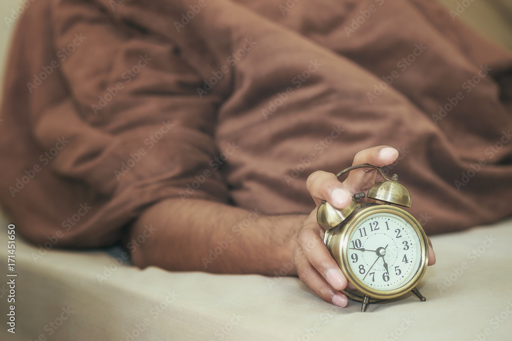 A man lying under a brown blanket is going off the alarm clock with drowsiness. A man lying under a brown blanket is going off the alarm clock with drowsiness.