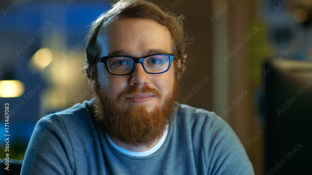 Young Bearded Creative Man With Glasses Thinks on a Problem While Working on His Personal Computer. He Looks Smilingly Into the Camera. Young Bearded Creative Man With Glasses Thinks on a Problem While Working on His Personal Computer. He Looks Smilingly Into the Camera.