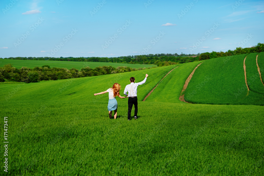 Young happy lovers running on meadow with green grass and blue sky, From the back Young happy lovers running on meadow with green grass and blue sky, From the back