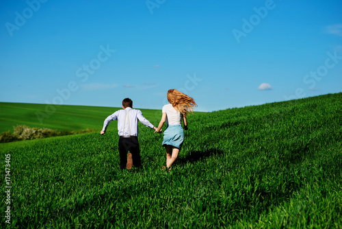 Young happy lovers running on meadow with green grass and blue sky, From the back